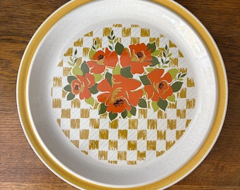 May include: Round ceramic serving platter with a white speckled surface and a yellow rim. The center features a floral design with orange flowers, green leaves, and a gold and white checkered pattern.