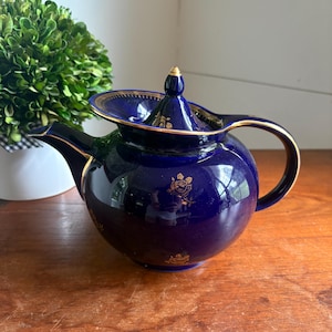 May include: A deep cobalt blue ceramic teapot featuring a rounded body, a curved handle, and a pointed lid. The teapot is decorated with delicate gold floral patterns and gold trim along the rim, spout, handle, and the edge of the lid.