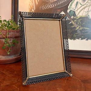 May include: A rectangular picture frame featuring a central gold-toned metal border surrounded by a dark, intricate, openwork metal lattice edge. The frame has decorative gold-toned corner accents and contains a plain, light brown backing board.