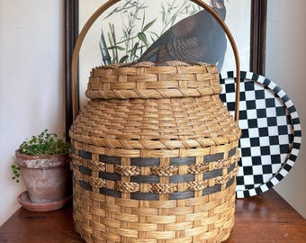May include: A cylindrical woven basket with a lid and a tall, arched wooden handle. The basket features light tan natural reed strips woven with horizontal bands of dark charcoal gray and braided tan accents around the center section.