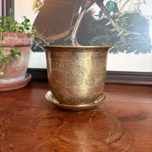 May include: A polished brass planter with a matching saucer, featuring intricate etched designs of birds and foliage around the exterior. The metal pot has a flared rim and a tapered body, showcasing a warm, metallic gold-toned finish.