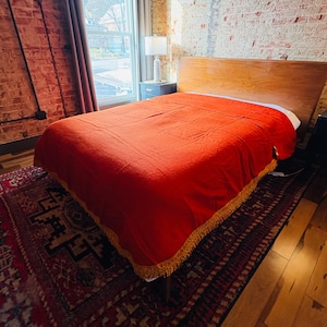 Op de afbeelding: Een bed met een houten hoofdbord is bedekt met een felrode deken. De deken heeft een gouden franje langs de onderrand. Onder het bed ligt een patroonkleed in rood- en zwarttinten.