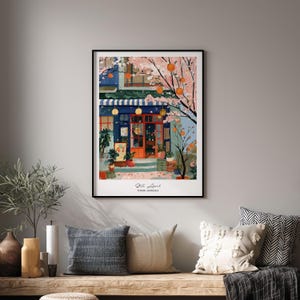 May include: A framed art print depicting a quaint shop with a blue facade, orange door, and striped awning. Cherry blossom trees with orange fruit frame the shop. The print is displayed above a wooden bench with pillows.
