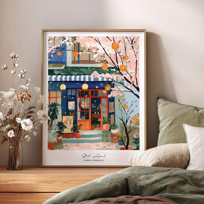 May include: A framed art print of a colorful shop scene. The shop has a blue awning, orange door, and is surrounded by flowering trees with orange fruit. The text "Still April, Tender Addresses" is at the bottom.