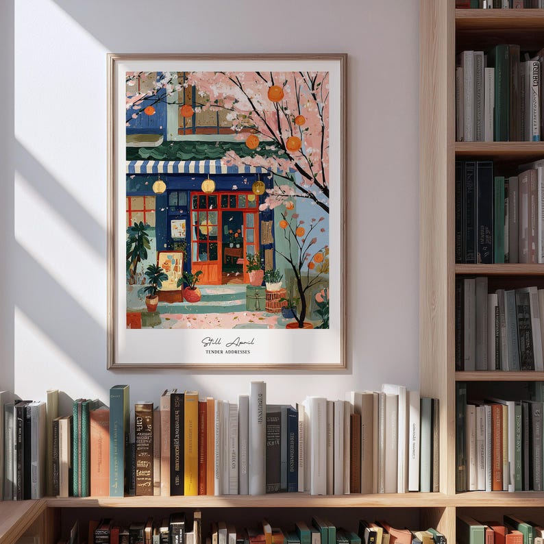 May include: A framed art print depicts a colorful street scene with a blue building, orange door, and a striped awning. Cherry blossom branches and orange fruit trees frame the building. The print is titled "Still April" and is displayed on a bookshelf.