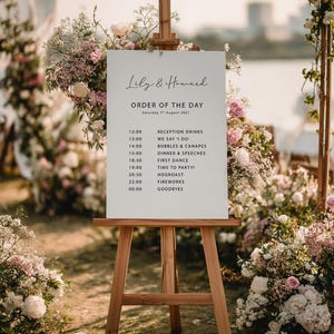 May include: A wooden easel displays a white sign with the text "Lily & Howard" and "Order of the Day" for a wedding. The schedule includes reception drinks, dinner, speeches, and fireworks. The sign is surrounded by floral arrangements.