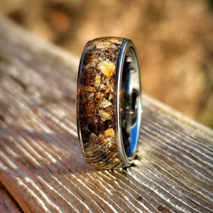 May include: A close-up of a ring with a silver band and a clear resin inlay filled with a mix of brown, tan, and beige organic materials. The ring is set against a blurred wooden background.
