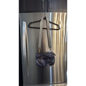 May include: A small, dark blue crossbody bag with a floral pattern hangs from a black hanger. The bag has a black zipper and a striped strap with gold-colored hardware. The bag is hanging on a stainless steel refrigerator.