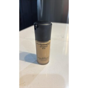 May include: A cylindrical glass bottle of MAC Studio Fix Fluid Broad Spectrum SPF 15 foundation with a black pump cap. The beige liquid makeup is visible through the clear glass, and the label features black text detailing the product name.