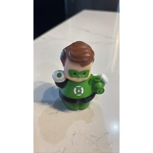 May include: A Green Lantern action figure with a green and black costume, brown hair, and a green mask. The figure is holding a green lantern and a white and green object.