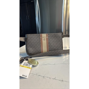 May include: A brown and tan Michael Kors fanny pack with a repeating MK logo pattern. The bag has a vertical stripe in the center with a tan and pink stripe. The bag has a gold-tone metal charm and a tag that says "Michael Kors".