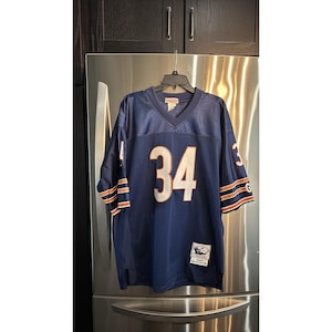 May include: A navy blue football jersey with the number 34 in white on the front and back. The sleeves have orange and white stripes. The jersey is hanging on a black hanger.