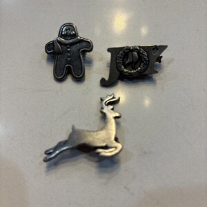 May include: Three silver-toned Christmas pins. One pin is a gingerbread man, another says "JOY" with a wreath, and the third is a reindeer. The pins are likely made of metal and are designed to be attached to clothing or accessories.