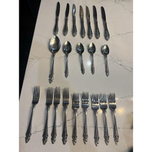 May include: A set of vintage silverware, including knives, spoons, and forks, arranged on a white surface. The silverware has a decorative floral pattern on the handles. The set includes various sizes of spoons and forks.