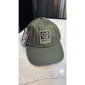 May include: Olive green baseball cap with a curved brim. The cap features a square patch with the word "DIXXON" in white letters, framed by a gold wreath and crossed axes. A black tag hangs from the cap.