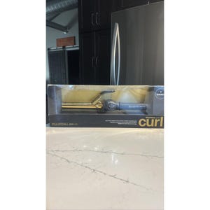 May include: A Paul Mitchell Express Gold curling iron in its original packaging. The curling iron has a gold barrel and a dark gray handle. The packaging is black with the words "express gold curl" printed on the front.