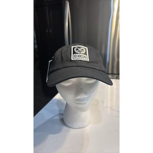 May include: Black baseball cap with a curved brim. The cap features a white square patch with the word "DIXXON" and a graphic design. The cap is displayed on a white mannequin head.