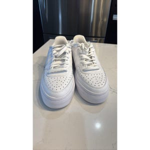 May include: Pair of white sneakers with a thick sole. The shoes have a perforated design on the toe box and white laces. The sneakers are made of a smooth material and are sitting on a white surface.