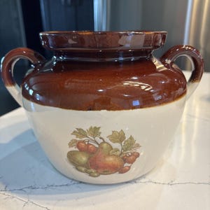 May include: A two-handled, ceramic crock pot with a brown top and handles, and a white base. The crock pot features a fruit and leaf design. The pot is suitable for cooking and serving.