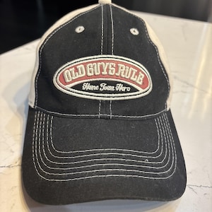 May include: Black and cream-colored baseball cap with the text "OLD GUYS RULE Home Town Hero" embroidered on the front. The cap has a curved brim and white stitching details.
