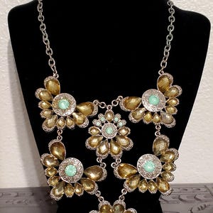 Op de afbeelding: Zilverkleurige statement ketting met grote, bloemvormige hangers met amberkleurige stenen en mintgroene middenaccenten. De ketting wordt getoond op een zwarte mannequin. Het label luidt "N-GN 117".