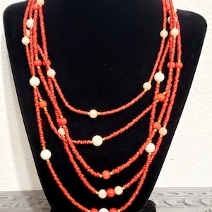 May include: A multi-strand necklace displayed on a black mannequin. The necklace features strands of small red beads interspersed with larger white and coral-colored beads. Gold-colored accents are visible. The tag reads "N-Rd 107".