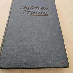 May include: A vintage cookbook titled "Kitchen Guide" with a blue cover. The title is in an elegant script. The book appears to be well-worn, with signs of age and use. The cover has a textured appearance.