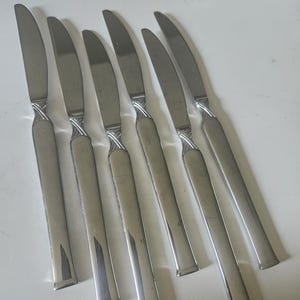 May include: A set of six silver-toned stainless steel dinner knives arranged in a row. Each knife features a smooth, flat handle and a curved blade, with a distinct decorative cross-hatched or braided metal texture detail located at the bolster.