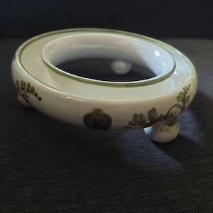 Pode incluir: Um suporte de anel de cerâmica branca com desenhos florais verdes e três pernas redondas. O suporte tem uma borda elevada ao redor da borda superior.