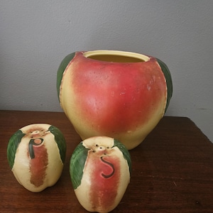 May include: A ceramic apple-shaped canister with a lid and two matching apple-shaped salt and pepper shakers. The canister and shakers feature a gradient of red, yellow, and green colors. The shakers are marked with the letters P and S in black.