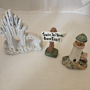 May include: Three ceramic figurines: a white wave, a sign that reads "Swim at your own risk!" and a white lighthouse with a green roof. The figurines are on a white surface.