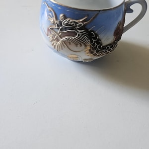 May include: A light blue porcelain teacup featuring a raised, three-dimensional dragon motif in black, white, and gold tones. The cup has a curved handle and a gold-trimmed rim, showcasing intricate dragon scales and a detailed head design.