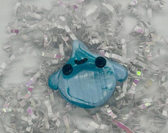 May include: A light blue, teardrop-shaped figurine with a smooth, glossy finish. The object features two dark, circular eyes and a small, curved mouth. The surface displays subtle white marbling and a slight shimmer throughout the material.