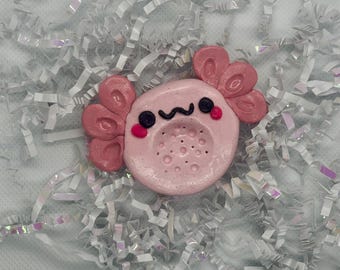 May include: A light pink axolotl-shaped object with a smooth, circular face, dark eyes, pink cheeks, and a black mouth. It features prominent, textured pink gills on both sides and a series of small, circular indentations on the lower face.