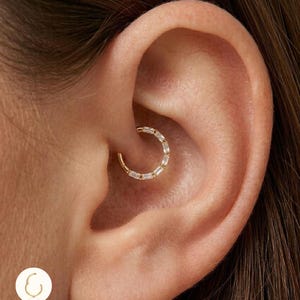 Peut inclure: Un bijou d'oreille à charnière de couleur or avec des pierres claires rectangulaires est montré dans un piercing à l'oreille. La boucle d'oreille est en titane G23. Le texte "G23 Titanium Hinged Clicker Hoop" est en bas à gauche.