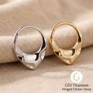 Puede incluir: Dos aros articulados, uno plateado y otro dorado, con un diseño texturizado en forma de lágrima. Los aros están hechos de titanio G23. El texto de la imagen dice "G23 Titanium Hinged Clicker Hoop."