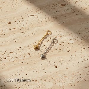 Op de afbeelding: Twee stervormige oorhangers, één goudkleurig en één zilverkleurig, elk hangend aan een ketting. De oorbellen worden getoond op een getextureerd, lichtgekleurd oppervlak. De tekst "G23 Titanium" is zichtbaar.