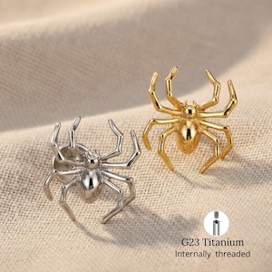 Pode incluir: Dois broches em forma de aranha, um prateado e outro dourado, são exibidos em uma superfície de tom neutro. Os broches têm pernas e corpos detalhados. O texto "G23 Titanium Internally threaded" é visível.