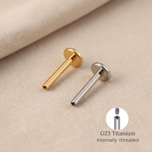 Może przedstawiać: Dwa labrety G23 z tytanu z gwintem wewnętrznym. Jeden jest w kolorze złotym, drugi srebrnym. Każdy labret ma płaską, okrągłą górę i prosty pręt z pustym końcem. Labrety są prezentowane na beżowej tkaninie.