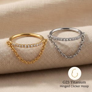 May include: Two hinged clicker hoops, one gold and one silver, each featuring a row of small, clear stones. A delicate chain hangs from each hoop. The text reads "G23 Titanium Hinged Clicker Hoop".