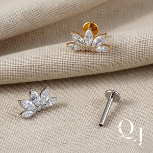 Puede incluir: Tres piezas de joyería: un pendiente de botón de flor de loto dorado y plateado, y un labret plateado. Los pendientes de botón de flor de loto están adornados con cristales transparentes y brillantes. El labret tiene una parte superior plana y redonda.