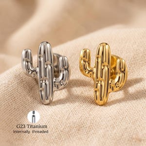 May include: Two cactus-shaped stud earrings, one silver and one gold, are shown on a beige surface. The earrings are made of G23 Titanium and are internally threaded.