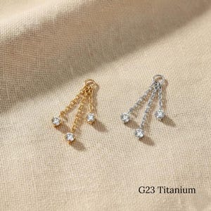 Könnte beinhalten: Zwei hängende Ohrringe, einer goldfarben und einer silberfarben, jeweils mit drei klaren, runden Edelsteinen. Die Ohrringe sind durch filigrane Ketten verbunden. Der Text "G23 Titanium" ist sichtbar.