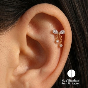 May include: A gold-toned titanium push pin labret earring featuring a bow-shaped design adorned with clear pear-cut and round-cut gemstones. Two delicate chains hang from the bow, each ending in a small, sparkling round-cut gemstone charm.