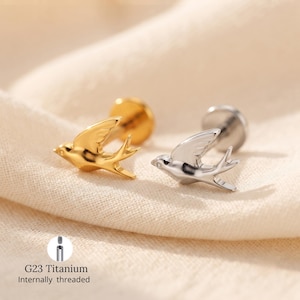 Könnte beinhalten: Zwei G23 Titan-Ohrstecker mit Innengewinde in Form von Schwalben. Ein Ohrstecker ist goldfarben, der andere silberfarben. Jeder Vogel zeigt detaillierte Flügel- und Schwanzschnitzereien und ist an einem flachen, runden Stift befestigt.