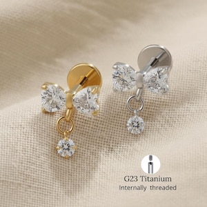 Pode incluir: Dois brincos em forma de laço, um dourado e outro prateado, cada um adornado com pedras preciosas transparentes e brilhantes. Uma pedra preciosa menor pende de cada laço. Os brincos são rotulados como "G23 Titanium Internally threaded".