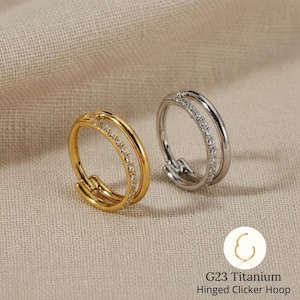 May include: Two hinged clicker hoop earrings, one gold-toned and one silver-toned, each with a double-band design. The inner band of each earring is adorned with small, sparkling stones. The text "G23 Titanium Hinged Clicker Hoop" is visible.