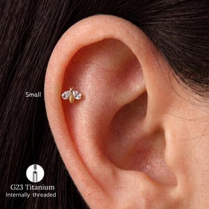 Könnte beinhalten: Ein goldfarbener Ohrstecker in Bienenform mit zwei klaren, tropfenförmigen Steinen als Flügel und einem strukturierten Körper. Das Schmuckstück ist als Small und G23 Titanium Internally threaded gekennzeichnet, inklusive einer Gewindedetailansicht.