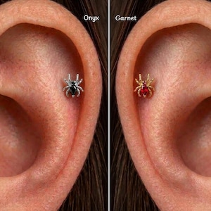 Könnte beinhalten: Zwei spinnenförmige Ohrpiercings. Das linke ist silberfarben mit einem schwarzen Onyx-Stein als Körper, beschriftet mit Onyx. Das rechte ist goldfarben mit einem roten Granat-Stein als Körper, beschriftet mit Garnet. Beide haben acht Beine und einen Edelstein.