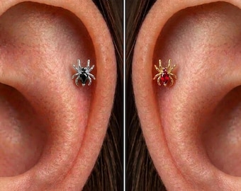 Puede incluir: Dos piercings de oreja con forma de araña. El de la izquierda es plateado con un cuerpo de piedra ónice negra, etiquetado como Onyx. El de la derecha es dorado con un cuerpo de piedra granate roja, etiquetado como Garnet. Ambos tienen ocho patas.
